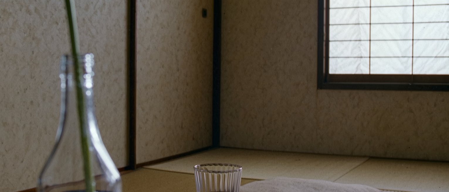 a low table seen close with a glass of water, a folded cloth, a single stem in a bottle and a tea bowl on its tray, two cushions pushed close against the table and the shoji screen close behind
