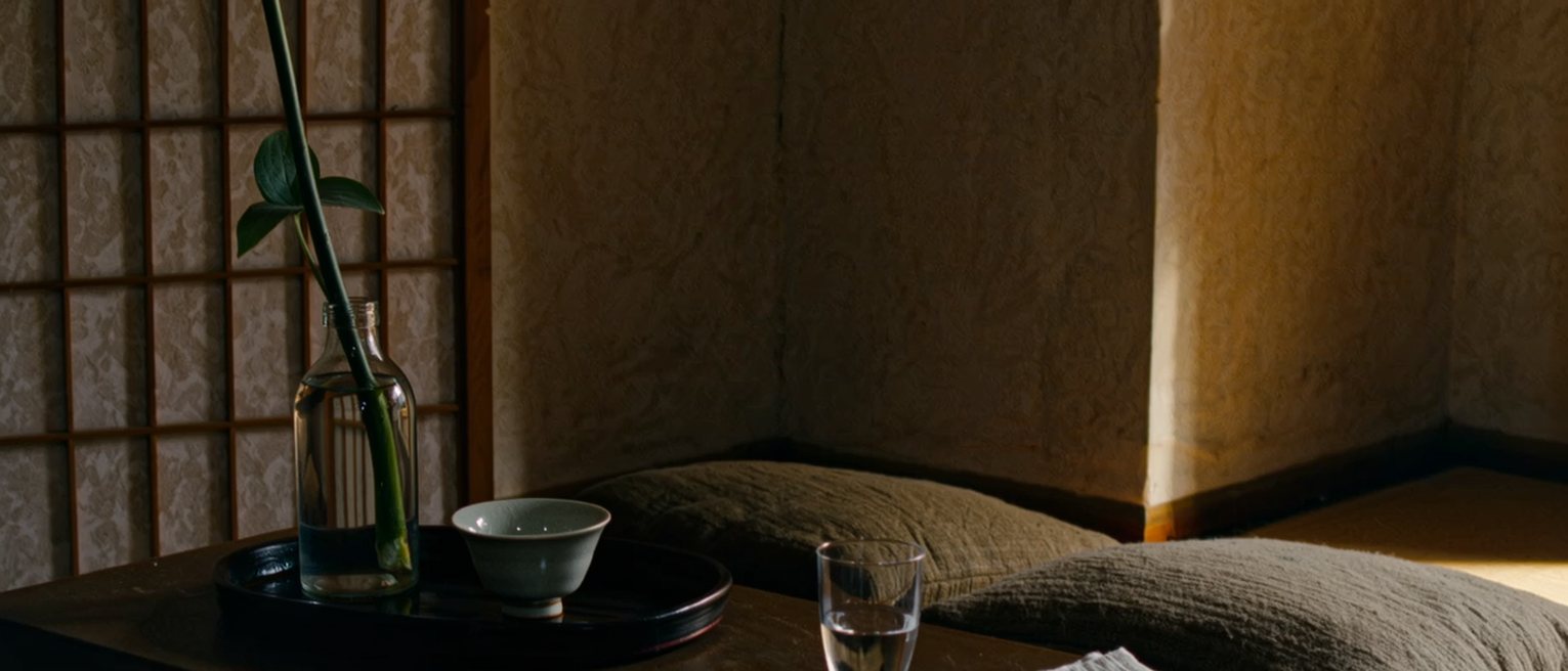 a low table seen close with a glass of water, a folded cloth, a single stem in a bottle and a tea bowl on its tray, two cushions pushed close against the table and the shoji screen close behind