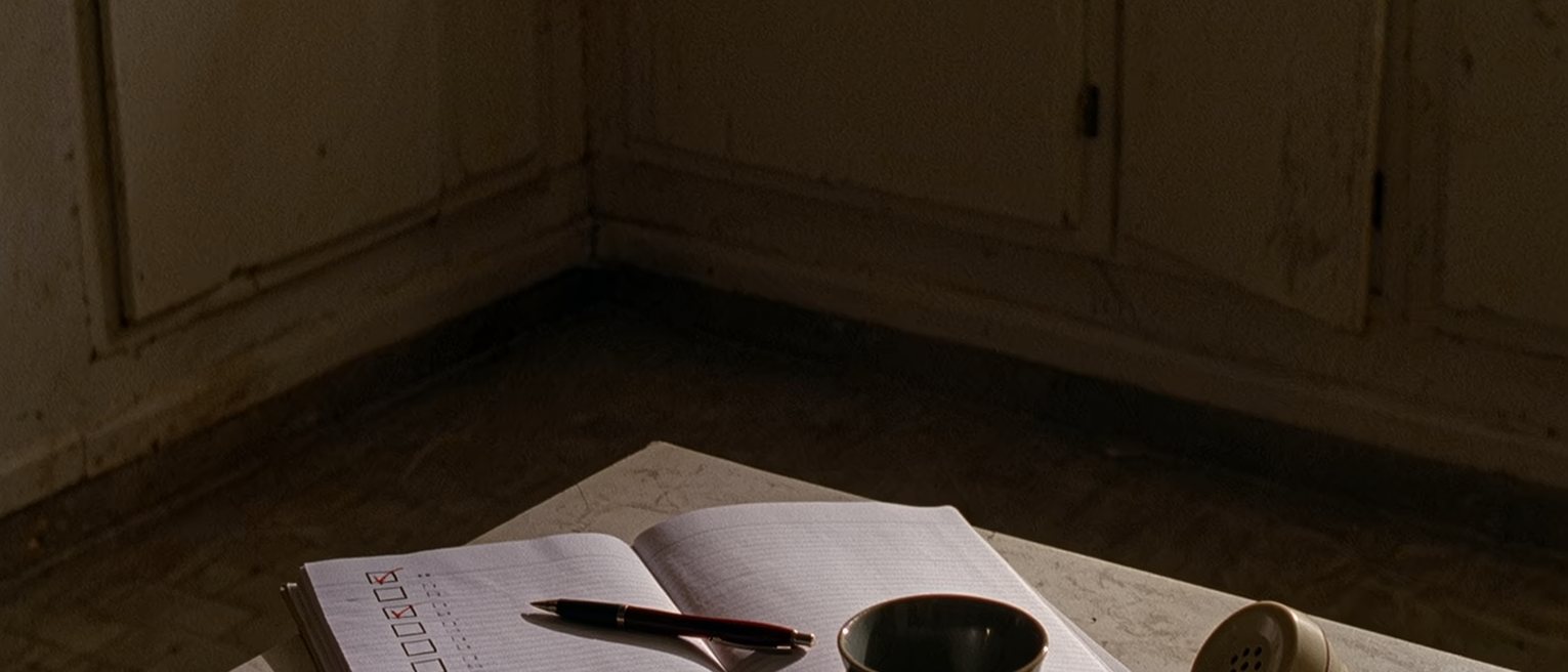 a kitchen table seen close with a lined notebook where a column of small boxes has been ticked in pencil, a pen laid across it, a cup, a handset face down at the edge, the window soft beyond