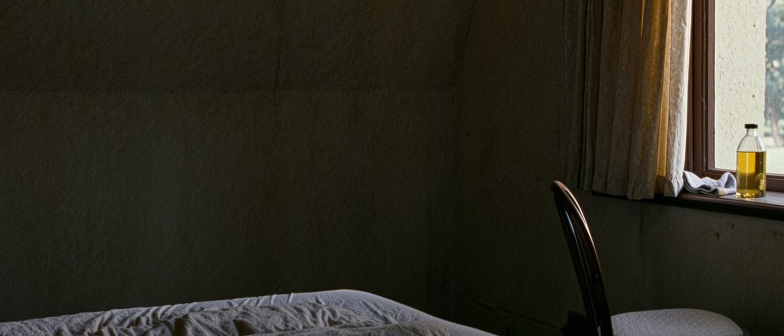 a bedroom with the curtains drawn and a single chair set facing the window, a bottle of oil and a folded cloth on the sill