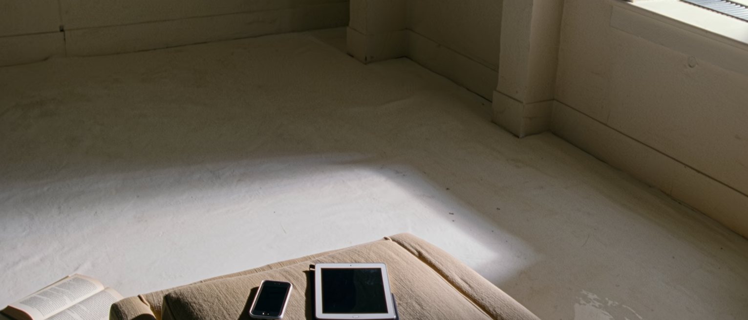 a low table in front of a sofa with a phone face down on it, a tablet face down beside the phone, and a book left open face down on the rug below, the sofa cushions still dented