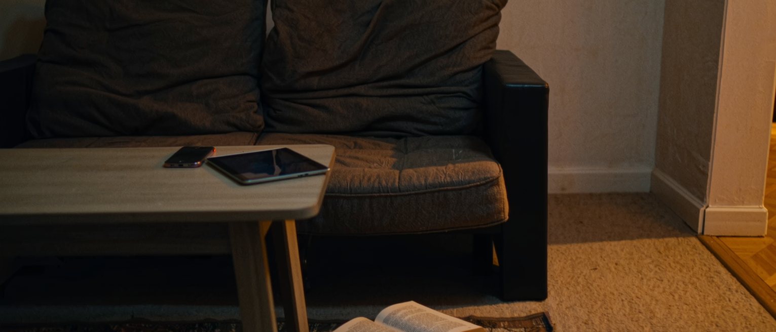 a low table in front of a sofa with a phone face down on it, a tablet face down beside the phone, and a book left open face down on the rug below, the sofa cushions still dented