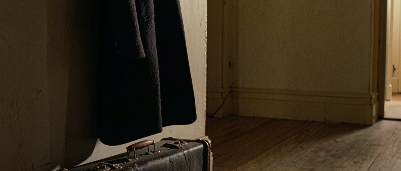 a hall with a coat on the hook and a suitcase beneath it that was packed once and never taken anywhere, dust on its handle