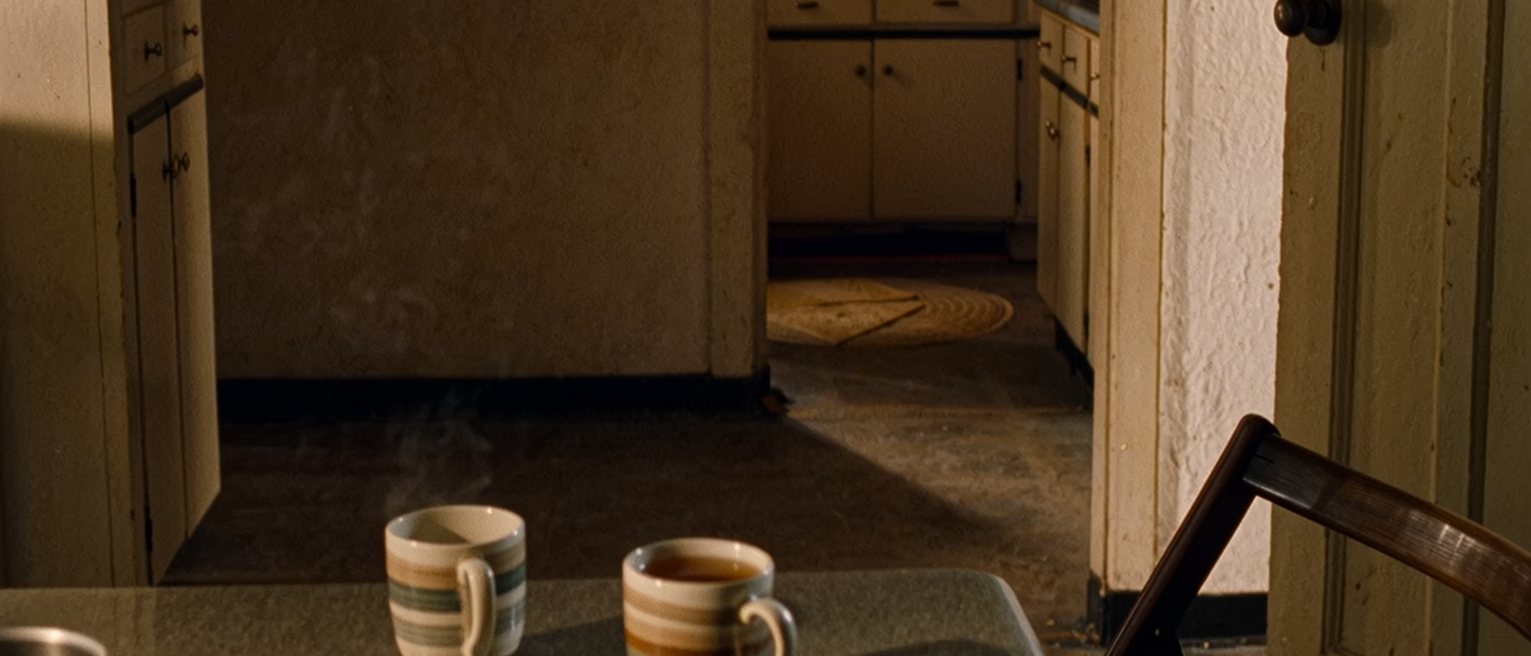 a kitchen with two cups set out and a pot of tea brewing, the second chair turned very slightly toward the door