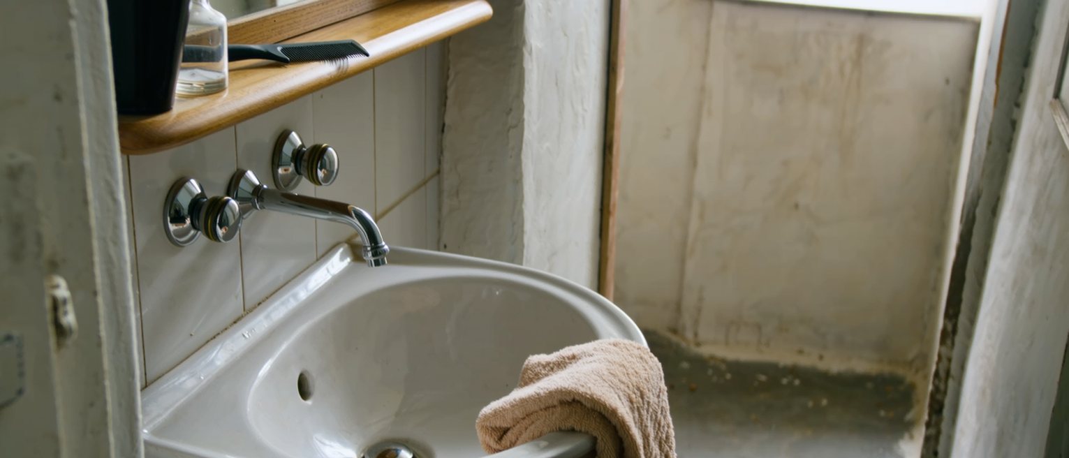 a bathroom seen close with a basin below the tap and a folded towel over its edge, a comb and a bottle with its cap off on the shelf above, the tiled wall soft beyond