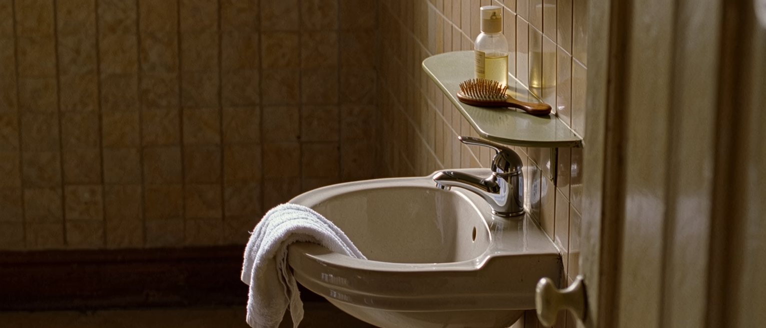 a bathroom seen close with a basin below the tap and a folded towel over its edge, a comb and a bottle with its cap off on the shelf above, the tiled wall soft beyond