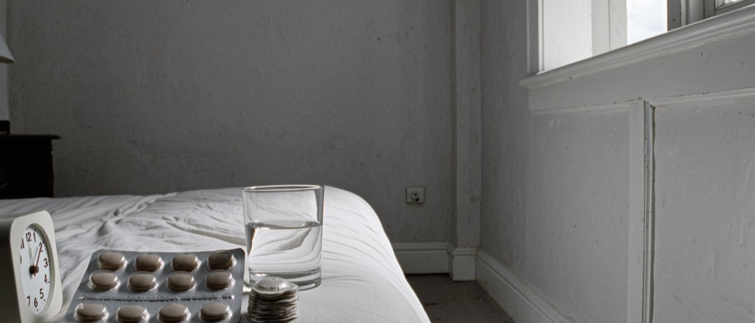 a bedroom seen close with an opened packet of adhesive patches and a small stack of used ones on the bedside table beside a glass of water and a clock, the bed turned back behind