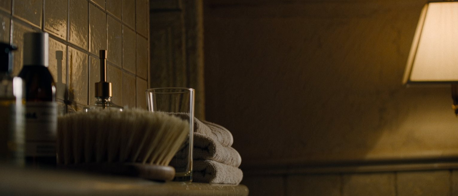 a bathroom shelf seen close with a soft bristle brush and a bottle of oil standing beside a folded towel and a glass, the tiled wall soft beyond