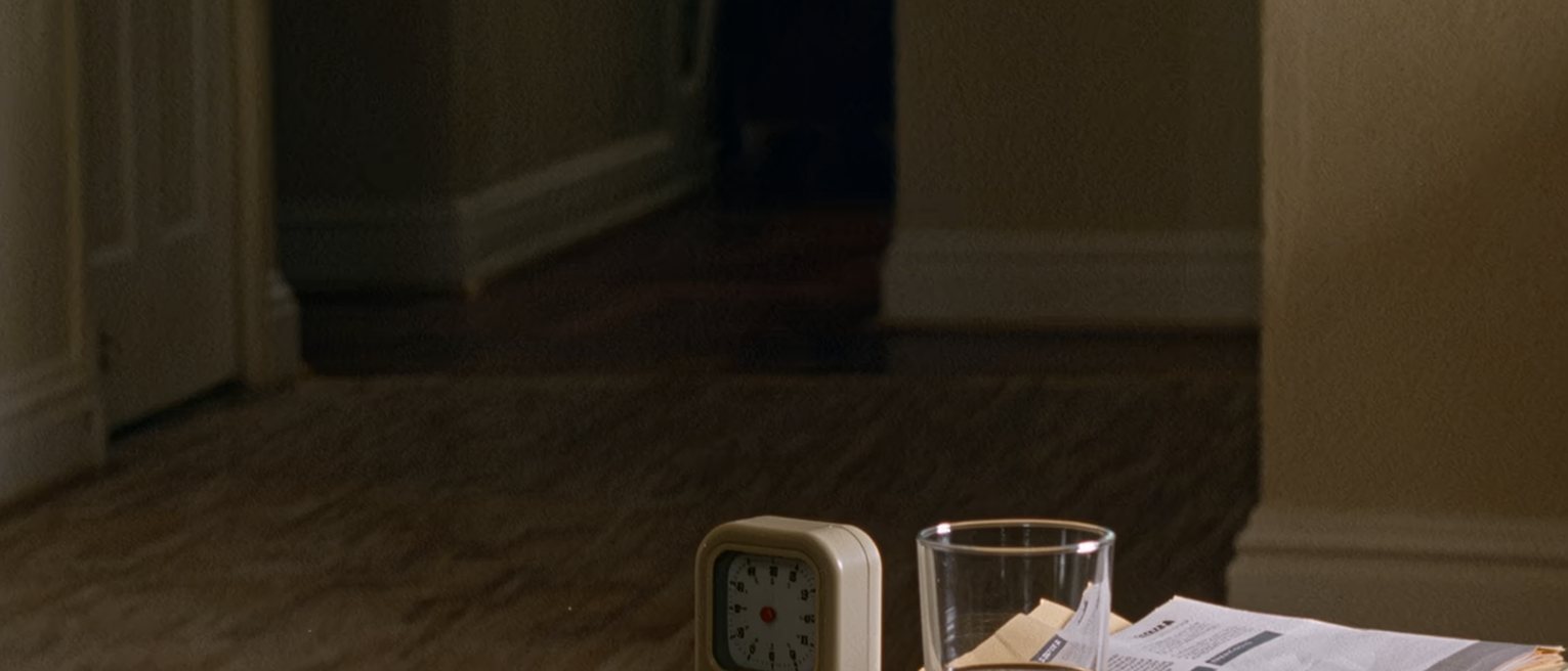 a table seen close with a small kitchen timer standing beside a glass of water and a folded printed sheet in a clear sleeve, a pen, the window soft beyond