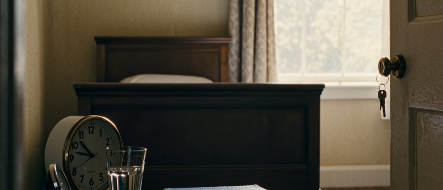 a bedroom seen close with a book face down on the cover, a glass of water and a small clock on the bedside table, the door closed with the key turned in it, the bedstead close behind and the curtains drawn beyond