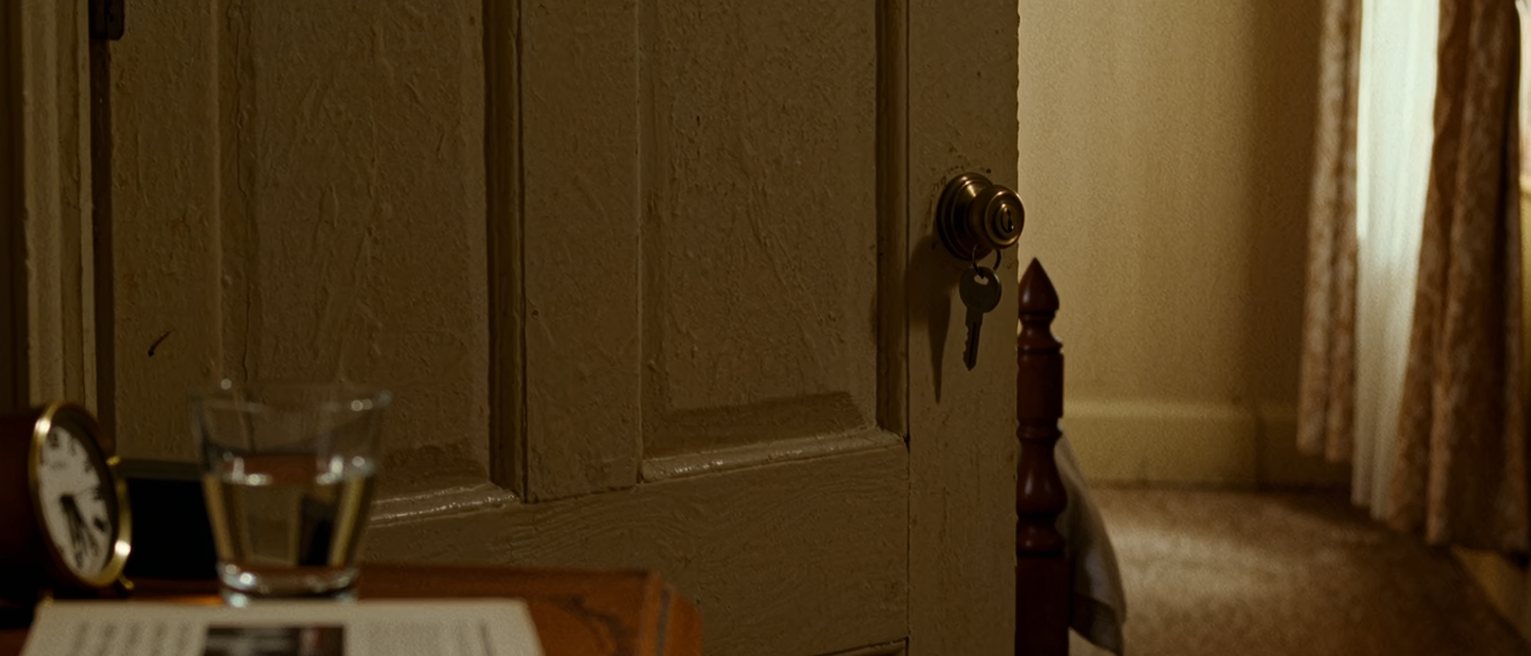 a bedroom seen close with a book face down on the cover, a glass of water and a small clock on the bedside table, the door closed with the key turned in it, the bedstead close behind and the curtains drawn beyond