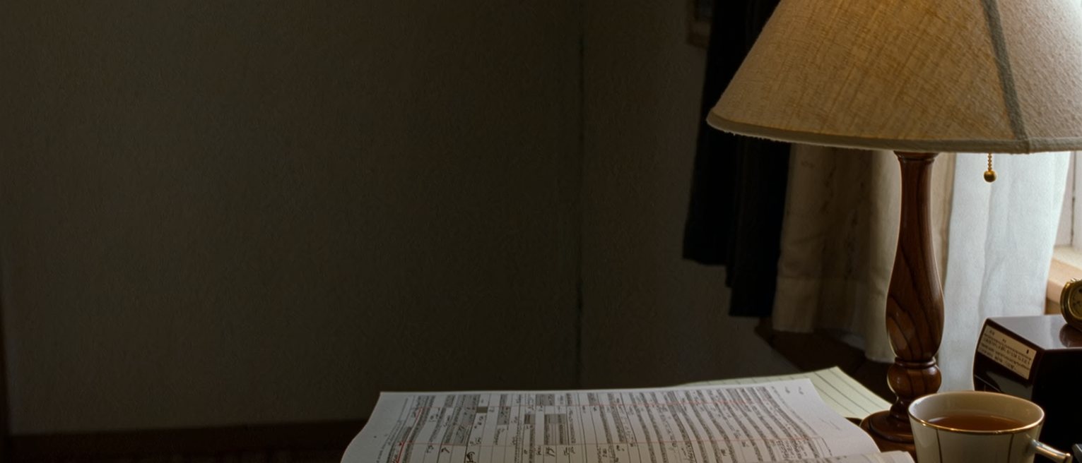 a writing desk seen close with a Japanese dictionary open at a page with one entry underlined in pencil, a notebook with a word written and crossed out, a cup of tea, the lamp close behind and the window soft beyond