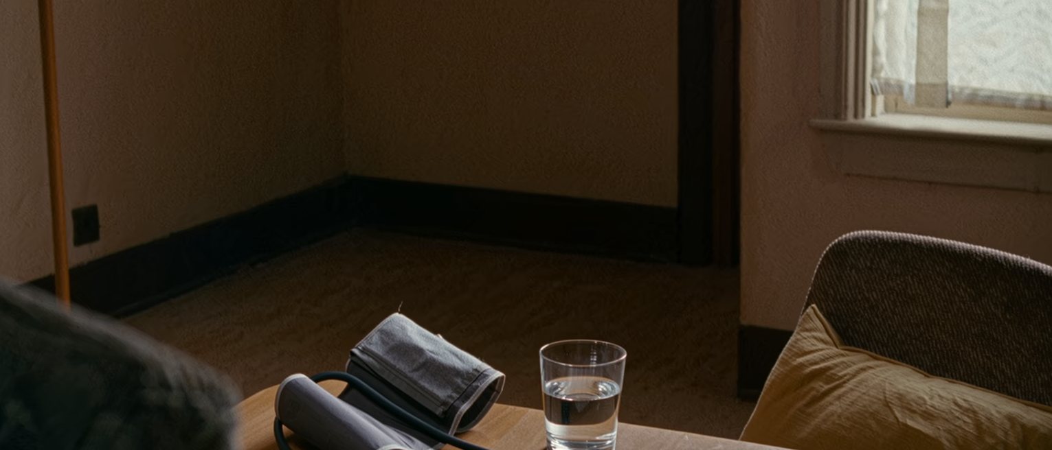a table seen close with a blood pressure cuff coiled beside a folded printout of readings and a pen, a glass of water, a cushion on the chair drawn up, the window soft beyond