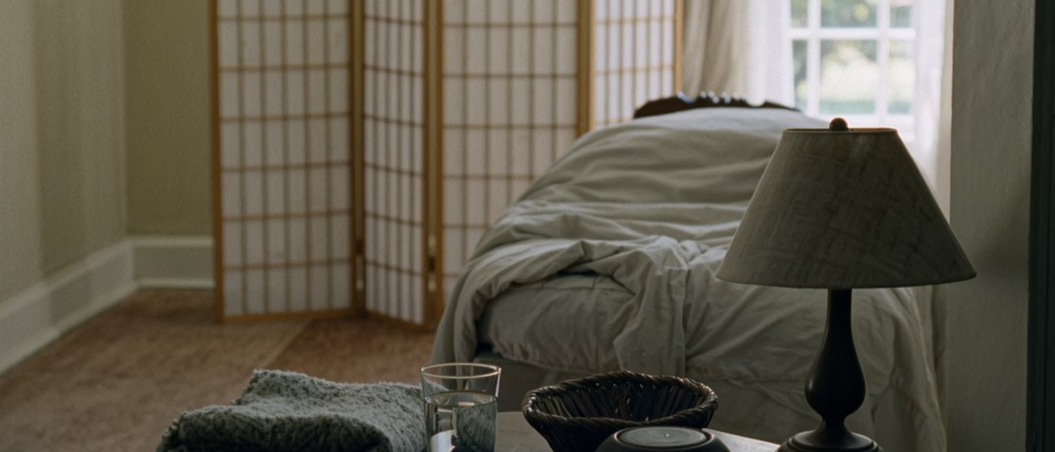 a quiet room seen close with a shaded lamp on a side table beside a folded blanket and a glass of water, the padded table with its sheet turned down close behind and the paper screen soft beyond