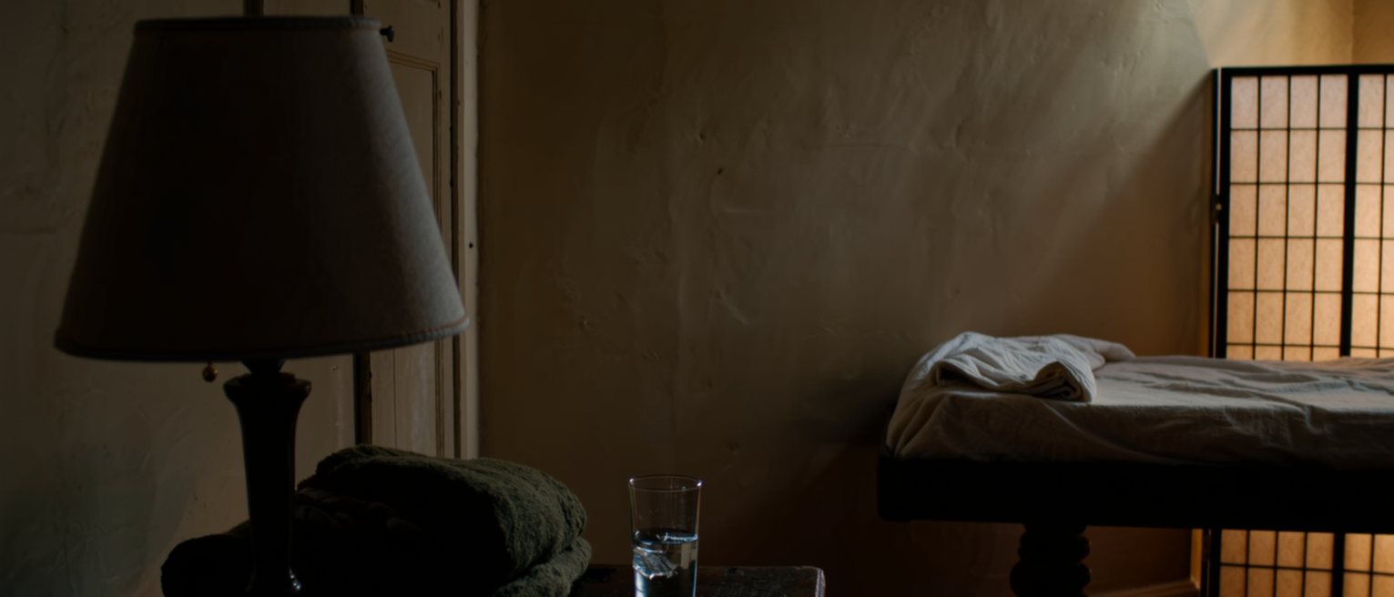 a quiet room seen close with a shaded lamp on a side table beside a folded blanket and a glass of water, the padded table with its sheet turned down close behind and the paper screen soft beyond