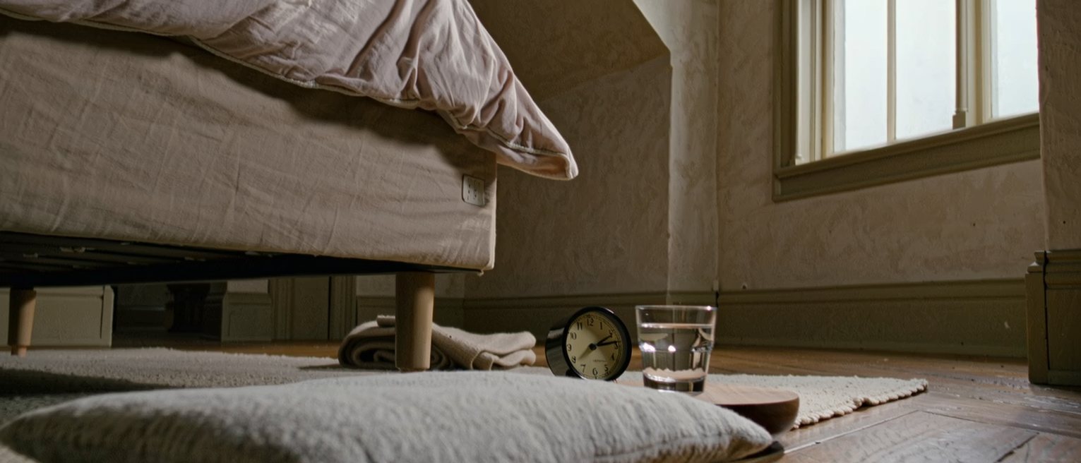 a bedroom seen close with a flat cushion squared on the rug beside the bed, a small clock and a glass of water set on the low stool above it, a folded blanket, the window soft beyond