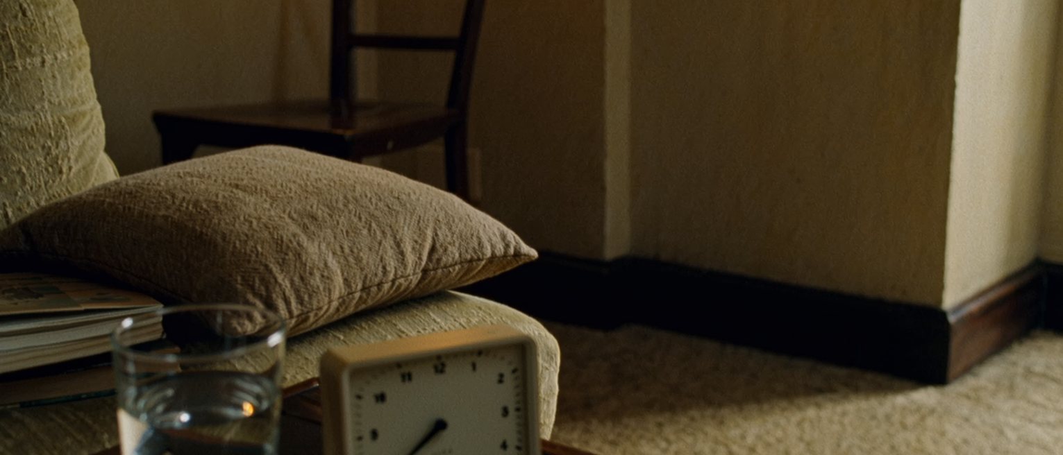 a sitting room seen close with a small timer standing beside a glass of water on the low table, a cushion set square on the chair behind them, a closed book, the room soft beyond