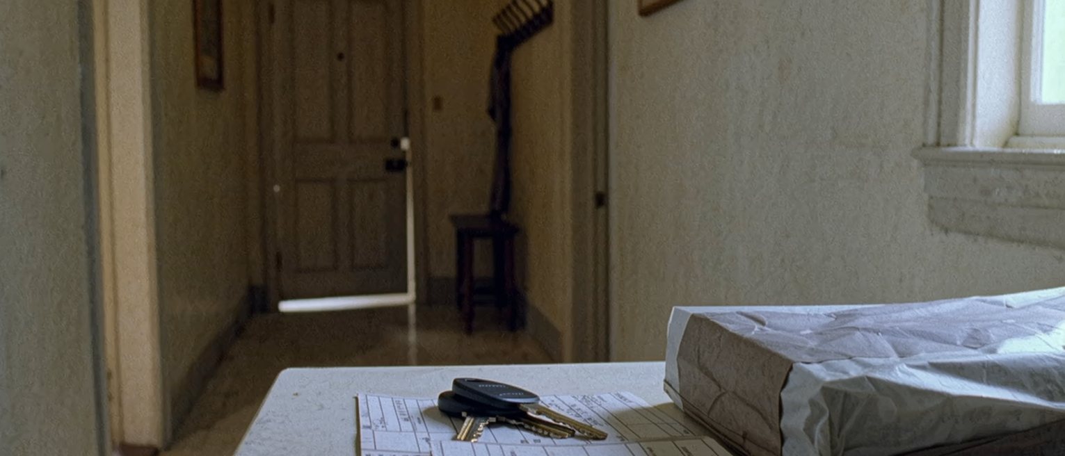 a hallway table seen close with an employee badge laid beside a set of keys, a folded shopping list and a lunch box packed and closed, the coat hooks close behind and the front door soft beyond