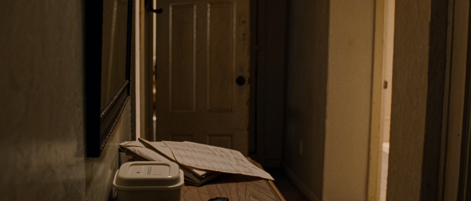 a hallway table seen close with an employee badge laid beside a set of keys, a folded shopping list and a lunch box packed and closed, the coat hooks close behind and the front door soft beyond