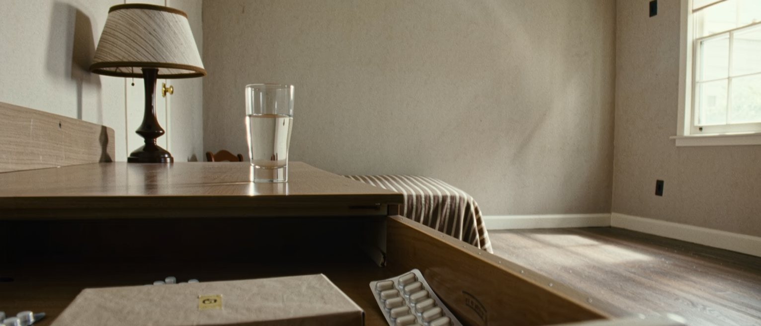 a bedside drawer seen close pulled open on a flat box still sealed and a strip of tablets in its packet, a glass of water and a lamp standing on the table above, the bed turned back behind