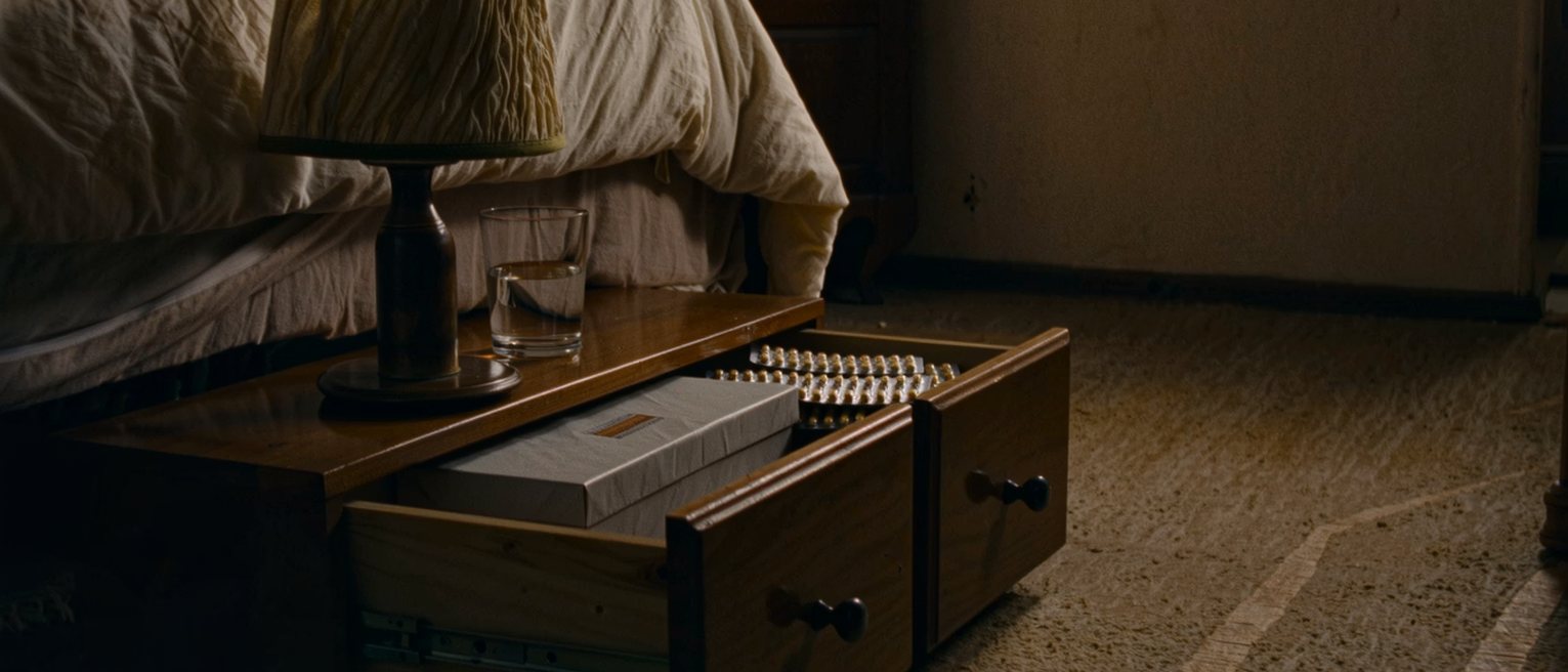 a bedside drawer seen close pulled open on a flat box still sealed and a strip of tablets in its packet, a glass of water and a lamp standing on the table above, the bed turned back behind