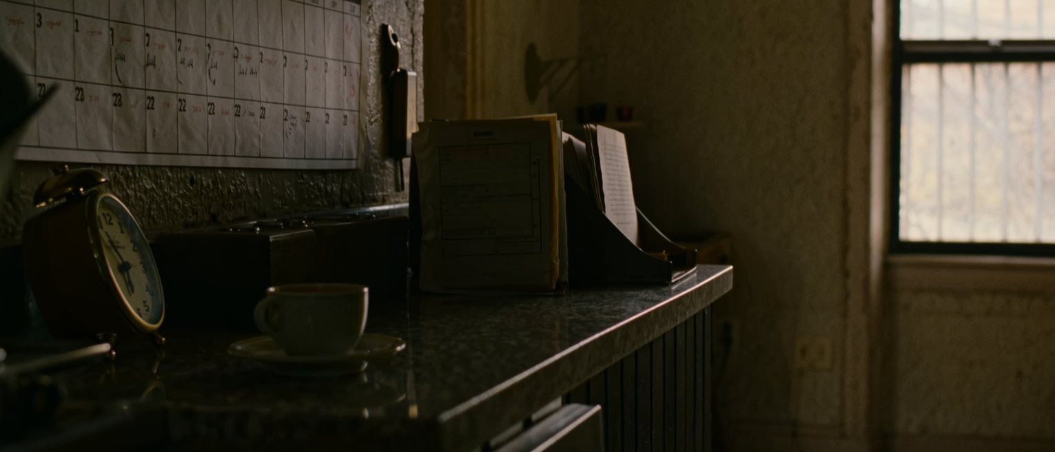 a kitchen seen close with a wall calendar where a run of days has been counted back in pencil, a cup of tea and a small clock on the counter beneath, a folder of papers propped at the side, the window soft beyond