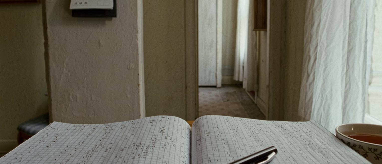a table seen close with a notebook lying open at a month of small pencil marks across its ruled squares, a pen laid across the page, a cup of tea, a wall calendar above, the window soft beyond