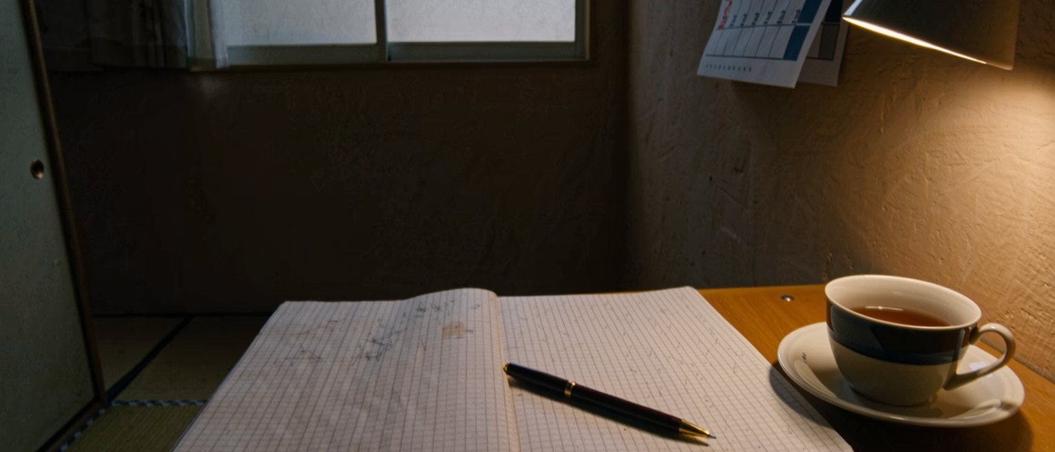a table seen close with a notebook lying open at a month of small pencil marks across its ruled squares, a pen laid across the page, a cup of tea, a wall calendar above, the window soft beyond