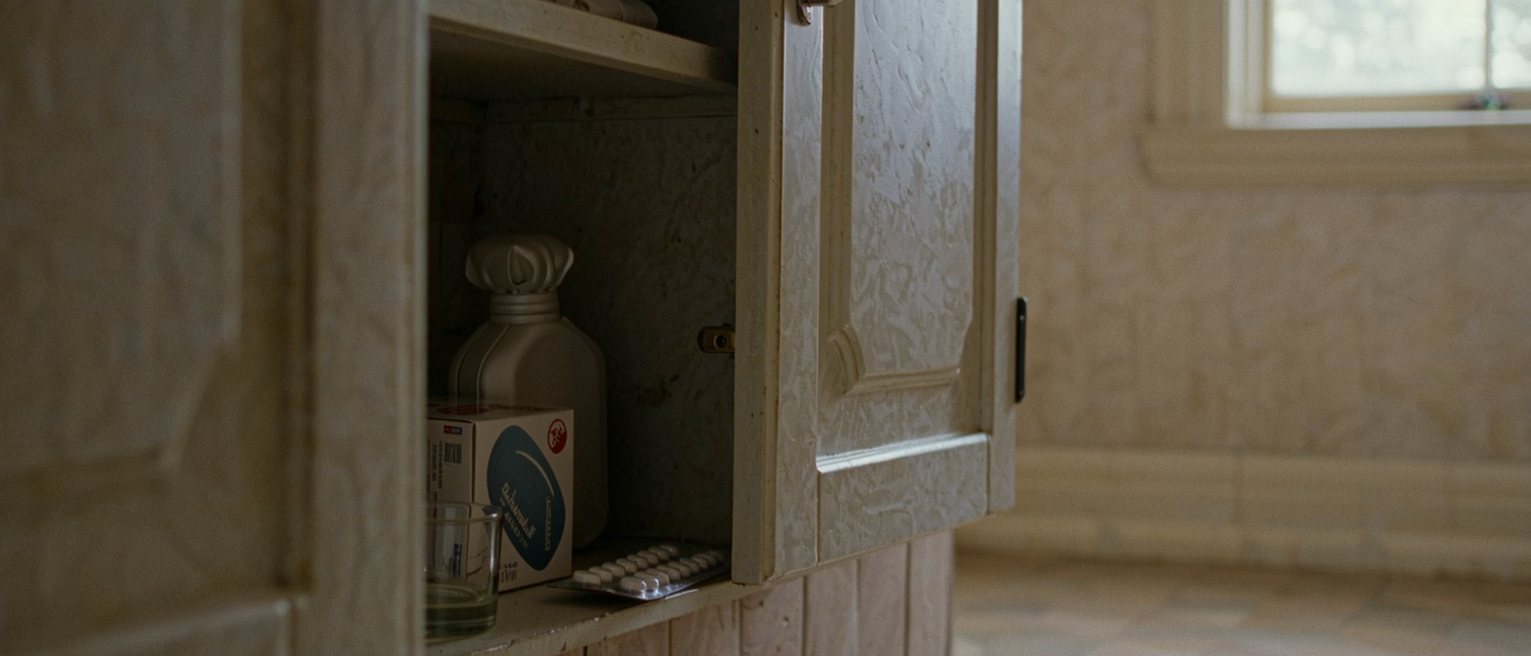 a bathroom cabinet seen close with its door open on a box of pads, a hot water bottle folded on the shelf below and a strip of tablets in its packet, a glass, the tiled wall soft beyond