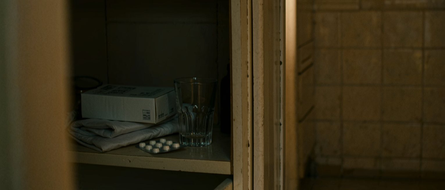 a bathroom cabinet seen close with its door open on a box of pads, a hot water bottle folded on the shelf below and a strip of tablets in its packet, a glass, the tiled wall soft beyond