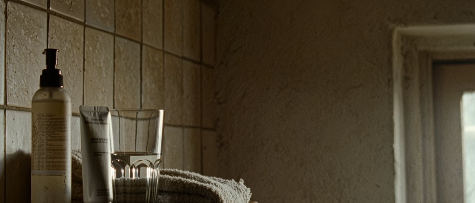 a bathroom shelf seen close with a prescription tube of cream beside a bottle of moisturiser, a folded flannel and a glass of water, the tiled wall close behind and the window soft beyond
