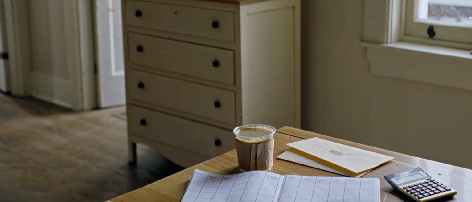 a kitchen table seen close with a weekly planner open with every hour filled in pen, a calculator, a cup of coffee gone cold and a folded envelope, the dresser close behind and the window soft beyond