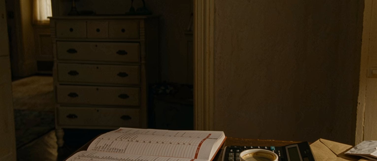 a kitchen table seen close with a weekly planner open with every hour filled in pen, a calculator, a cup of coffee gone cold and a folded envelope, the dresser close behind and the window soft beyond