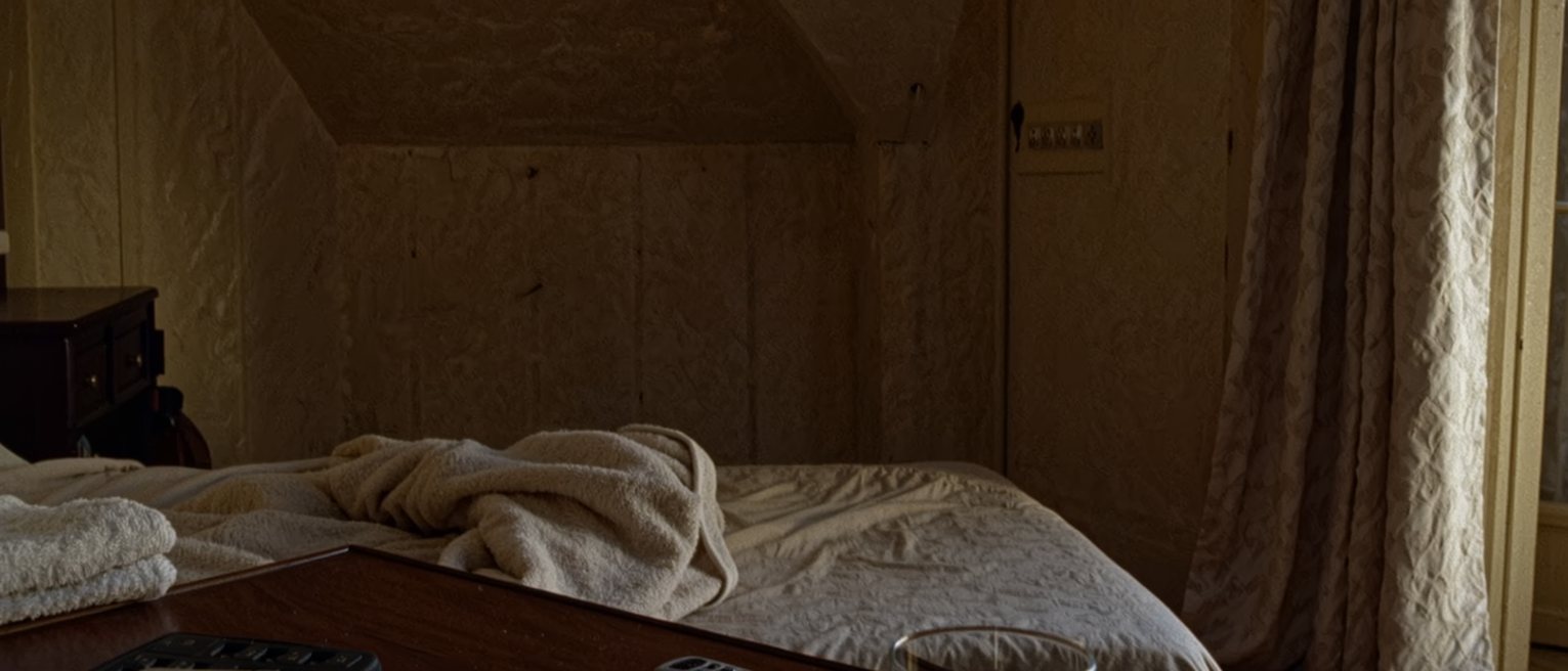 a bedside table seen close with a phone face down and switched off, a glass of water and a folded towel, the bed with its cover turned back close behind and the curtains drawn beyond