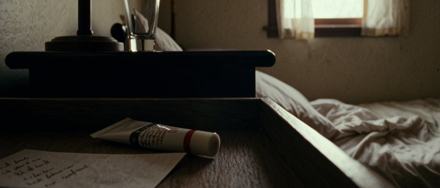a bedroom seen close with a bedside drawer open on a tube of gel still sealed and a folded card of notes, a lamp and a glass standing above, the bed turned back behind, the window soft beyond