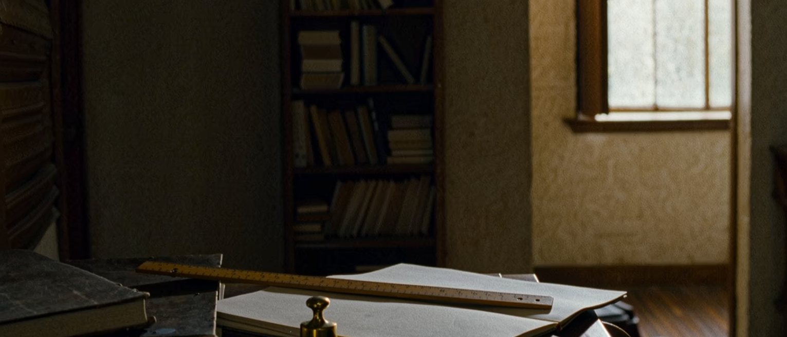 a study desk seen close with a wooden ruler laid across an open notebook, a small brass weight, a cup of tea and a pen, the bookshelf close behind and the window soft beyond