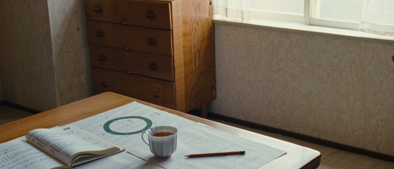 a kitchen table seen close with a printed four-circle diagram torn in half, a Japanese paperback open beside it, a pencil and a cup of tea, the dresser close behind and the window soft beyond