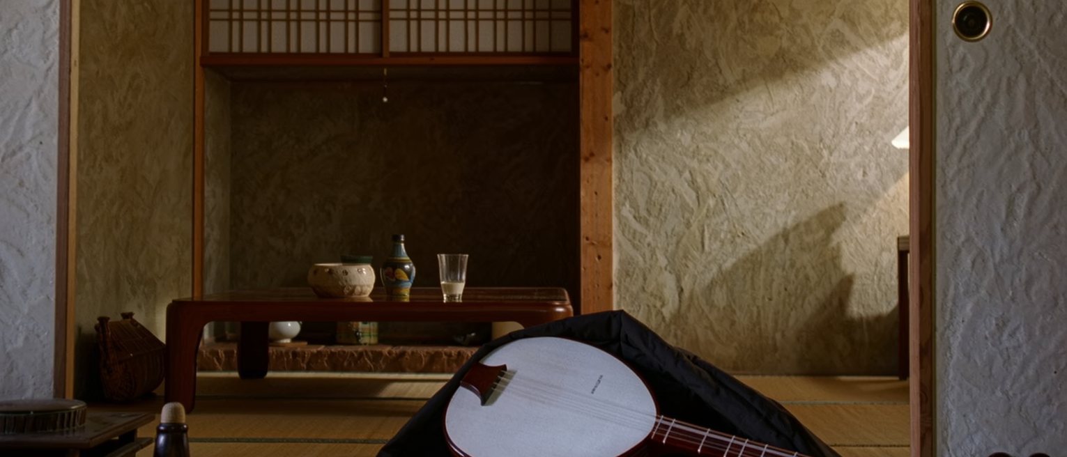 a tatami room seen close with a shamisen in its cloth cover laid on the floor beside a low table with a cup of sake untouched, the alcove close behind and the shoji soft beyond