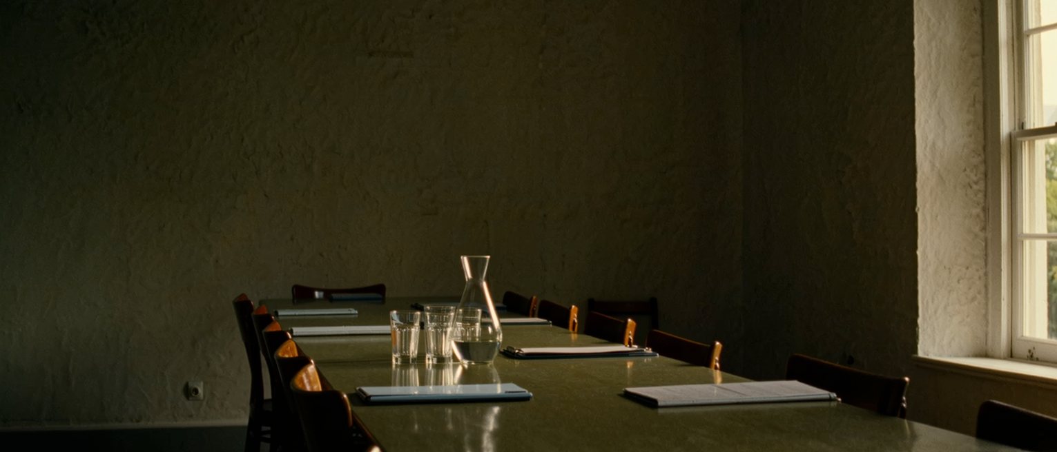 a long table seen close with a row of empty chairs down one side, a water jug and glasses, a document folder laid at each place, the far wall bare, the window soft beyond