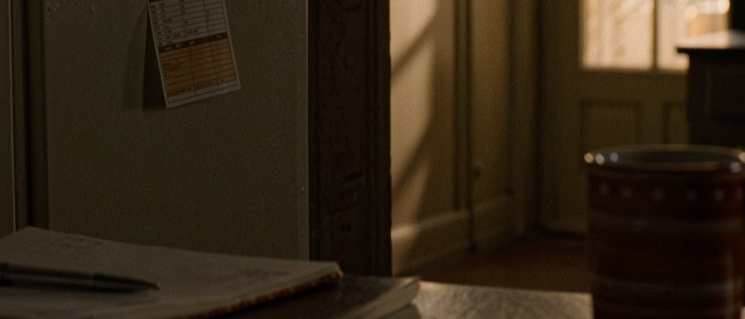a kitchen seen close with a small printed appointment card held under a magnet on the fridge door, a pen and a diary on the counter below it, a cup, the window soft beyond