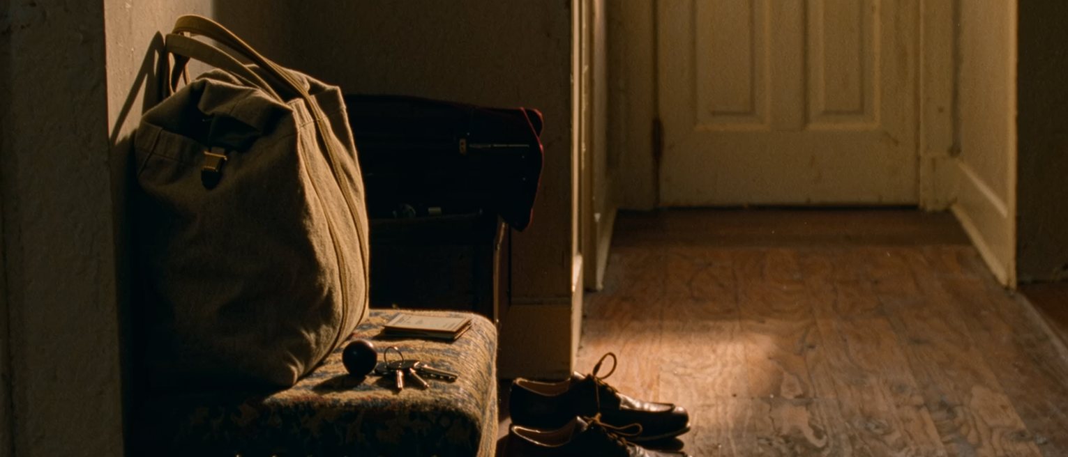 a hall seen close with a canvas bag packed and standing ready on the bench beside a folded card and a set of keys, a pair of low shoes below, the door soft beyond