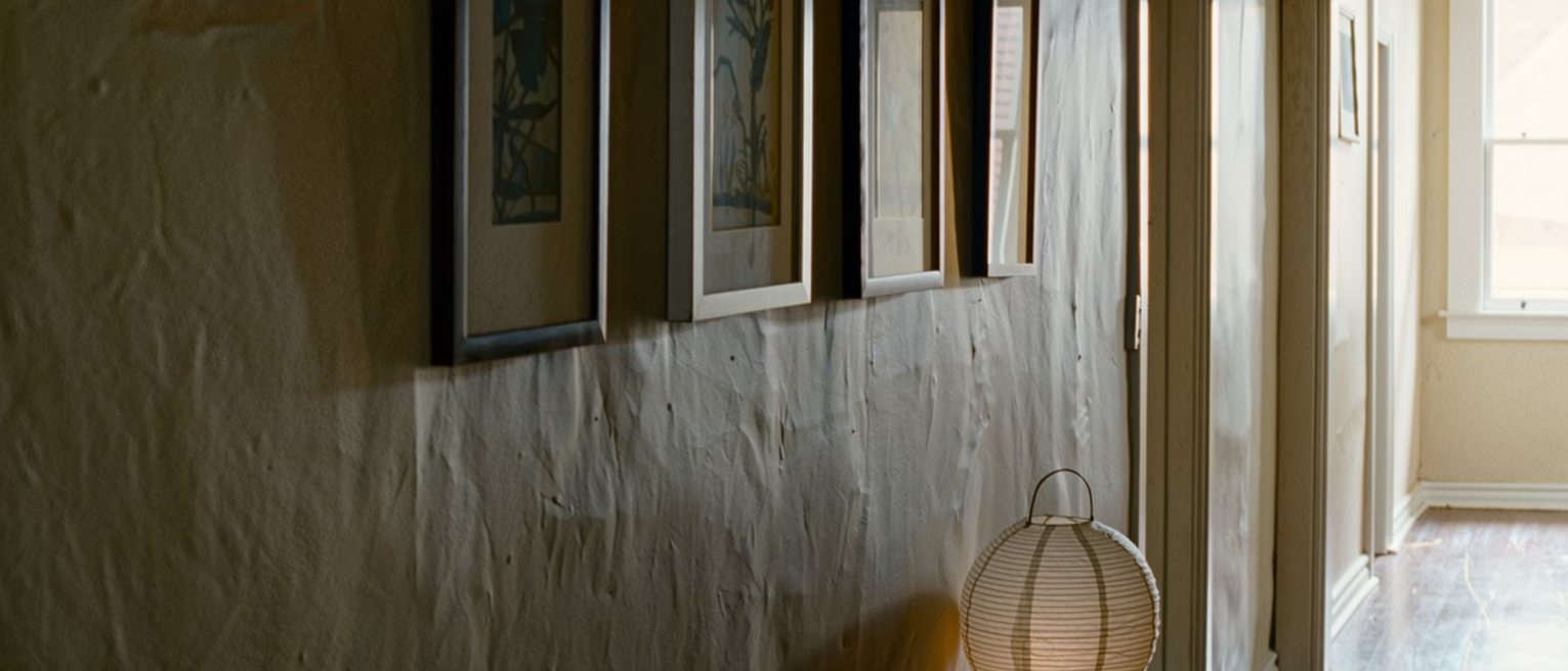 a hallway seen close with four framed prints hung on the wall each turned to face the wall, a low bench beneath them, a paper lantern on the bench, the corridor soft beyond
