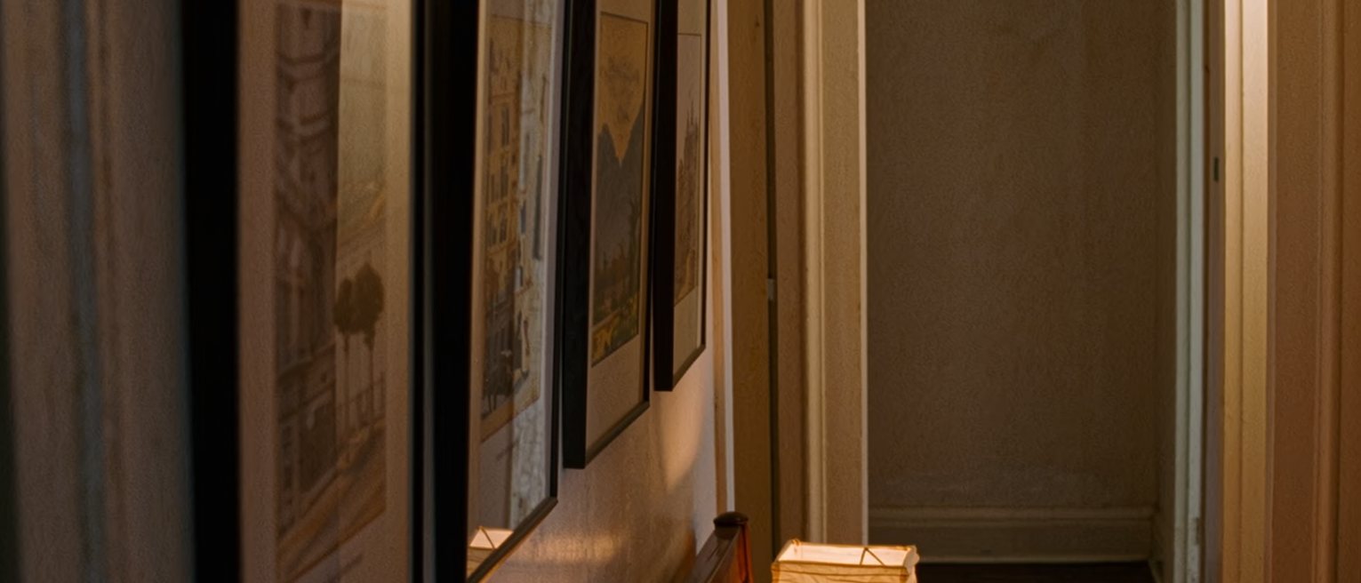 a hallway seen close with four framed prints hung on the wall each turned to face the wall, a low bench beneath them, a paper lantern on the bench, the corridor soft beyond