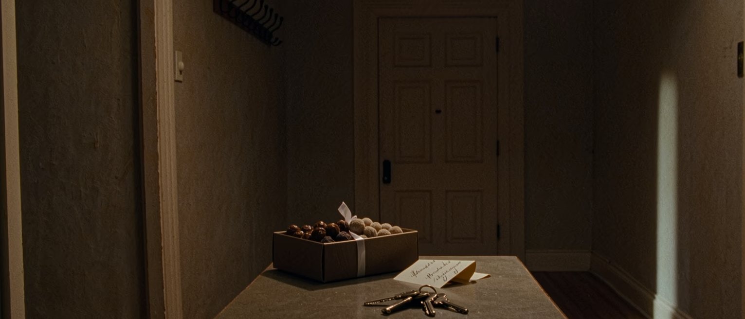 a hallway table seen close with a box of sweets with its ribbon untied, a folded handwritten thank-you note and a set of keys, the coat hooks close behind and the front door soft beyond