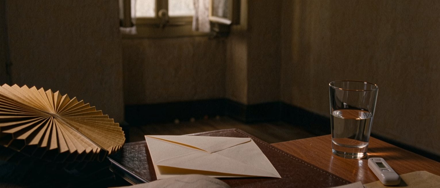 a desk seen close with a folded letter in its envelope, a paper fan, a glass of water beside a small thermometer, a diary open with several hours crossed out in pencil, the window soft beyond