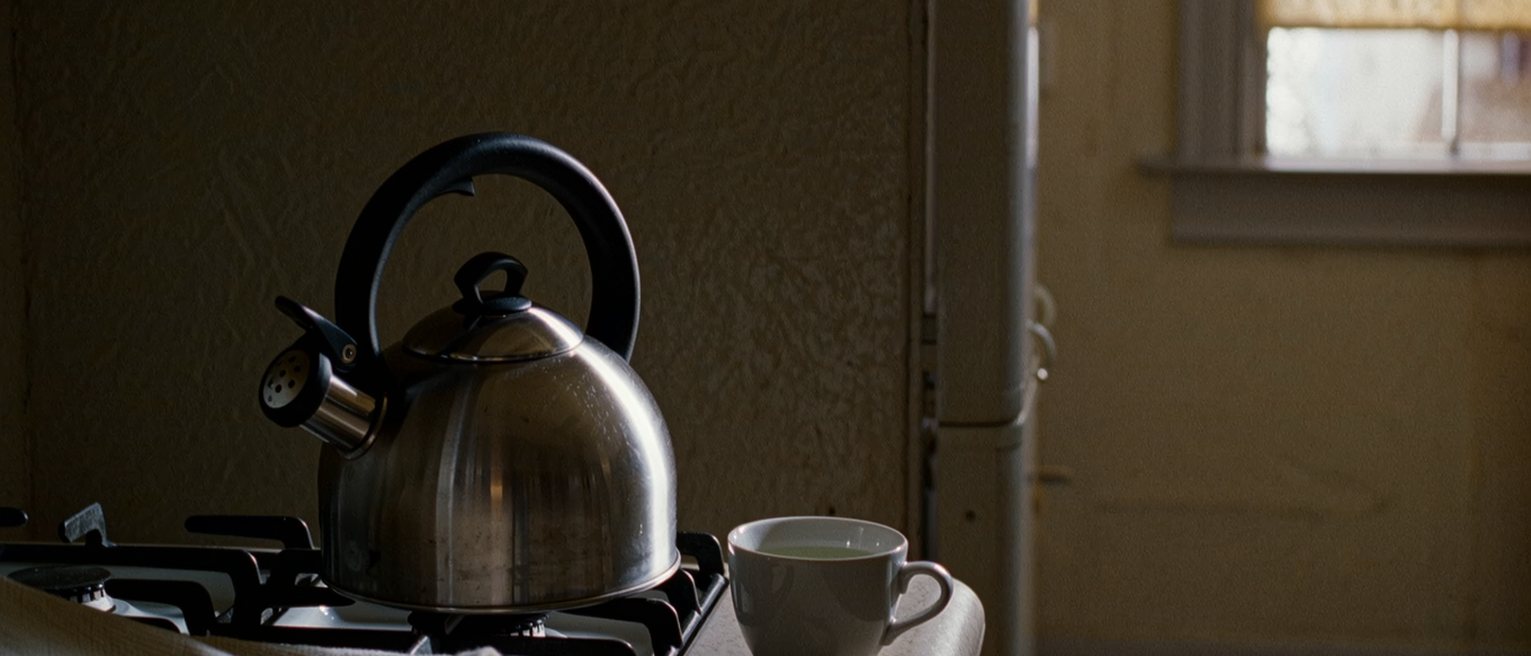 a kitchen seen close with a kettle lifted off the heat onto the cold ring beside it, the gas turned off, a cup of water and a folded cloth on the counter, the window soft beyond
