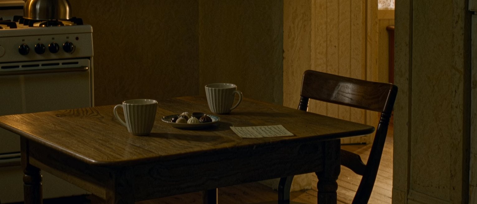 a kitchen table seen close with two cups set out and a plate of sweets, a second chair drawn up, a note left on the table, a kettle on the stove, the window soft beyond