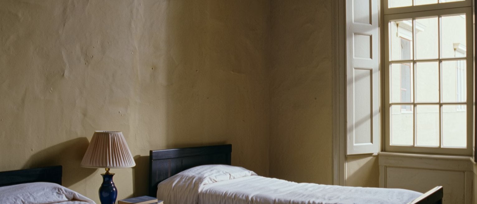 a bedroom seen close with two single beds set apart and a narrow table between them holding one lamp and two books, the covers turned back on both, a tall window soft beyond