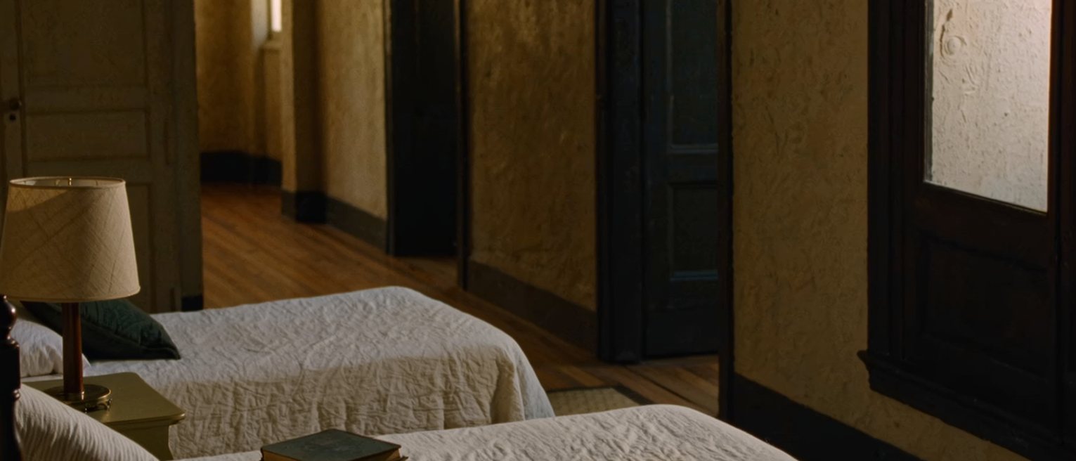 a bedroom seen close with two single beds set apart and a narrow table between them holding one lamp and two books, the covers turned back on both, a tall window soft beyond
