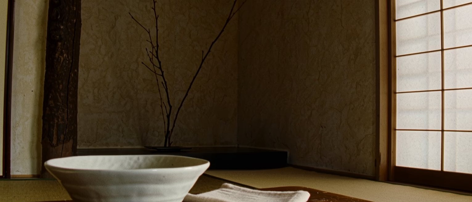 a tatami room seen close with a single tea bowl on a low table and a folded cloth beside it, the alcove with one branch in a vase close behind and the shoji soft beyond
